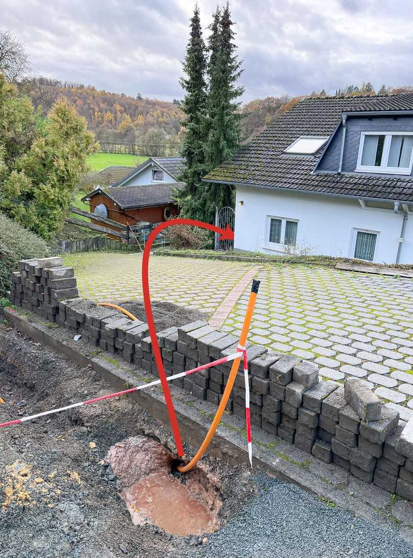 Blick auf das eingezogene Verbundrohr und die abschüssige Einfahrt des Grundstückes mit der Hausecke, um die herum in einem Winkel von 80° gebohrt werden musste. Der rote Pfeil markiert die Bohrrichtung. | Foto: L-Team X-Tools Blick auf das eingezogene Verbundrohr und die abschüssige Einfahrt des Grundstückes mit der Hausecke, um die herum in einem Winkel von 80° gebohrt werden musste. Der rote Pfeil markiert die Bohrrichtung. | Foto: L-Team X-Tools