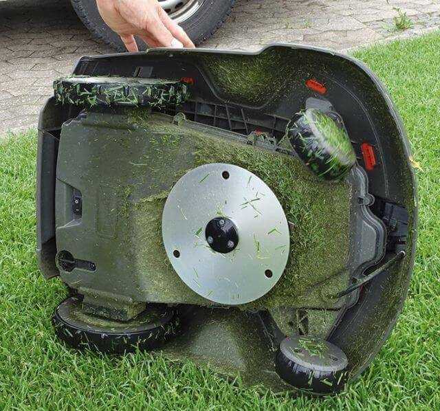 Automower von unten, die drei Messerklingen am Drehteller sind unsichtbar eingeklappt, die Verschmutzung mit Rasenpartikeln hält sich in Grenzen.